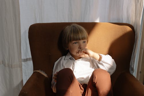 A Boy in White Striped Long Sleeves Sitting on a Brown Sofa