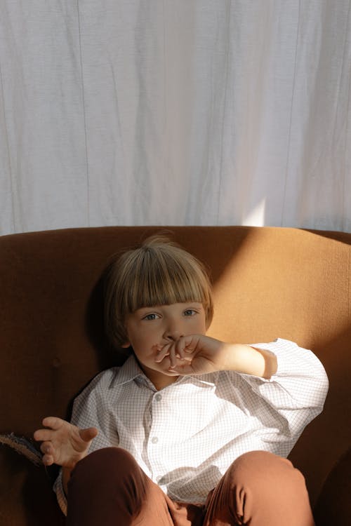 A Boy in White Striped Long Sleeves Sitting on a Brown Sofa