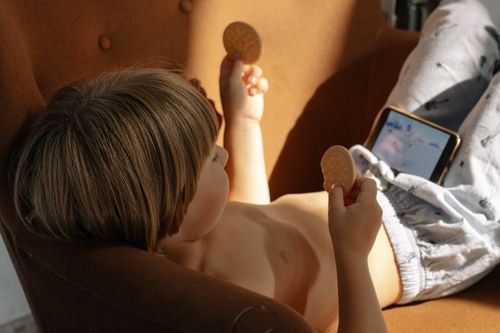 Free Young child lying on sofa holding snacks while watching a smartphone. Stock Photo