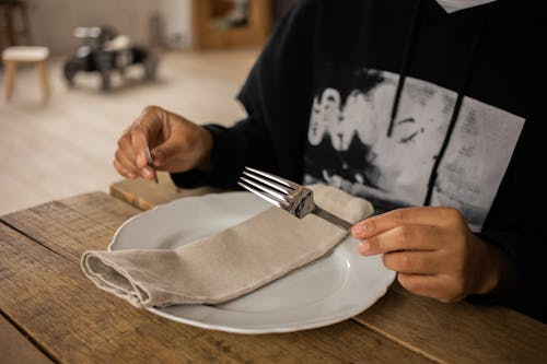 Crop anonymous person sitting at wooden table with fork and knife in hands near white plate with napkin on blurred background