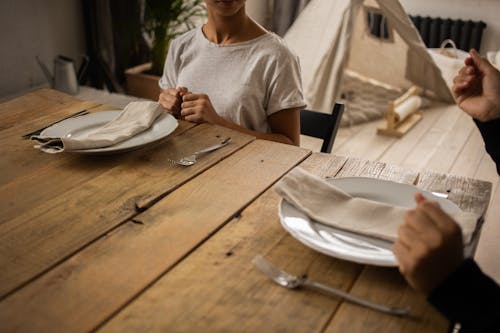 Crop faceless female sitting at wooden table with plate and cutlery near anonymous person while learning dining etiquette in light room