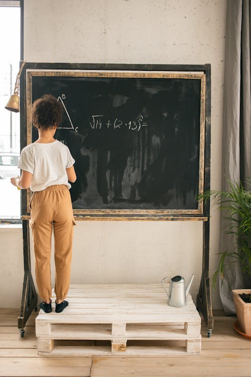 Back view full body of clever faceless girl writing on blackboard while solving mathematical equation during lesson in light room