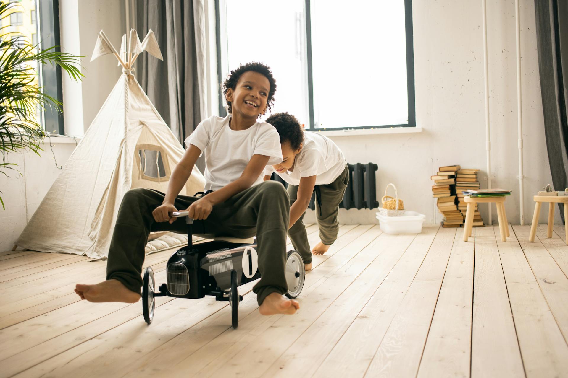 Happy barefoot African American brothers in casual wear riding toy car while playing together in light room with blanket fort