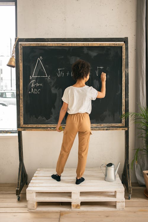 Full body back view of unrecognizable clever girl in casual clothes solving equation while writing on blackboard in light room