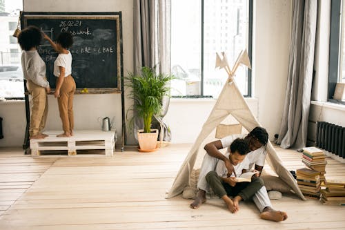 Full body of young African American man sitting on floor and helping little son to read book near wife and daughter writing on blackboard while doing homework assignment