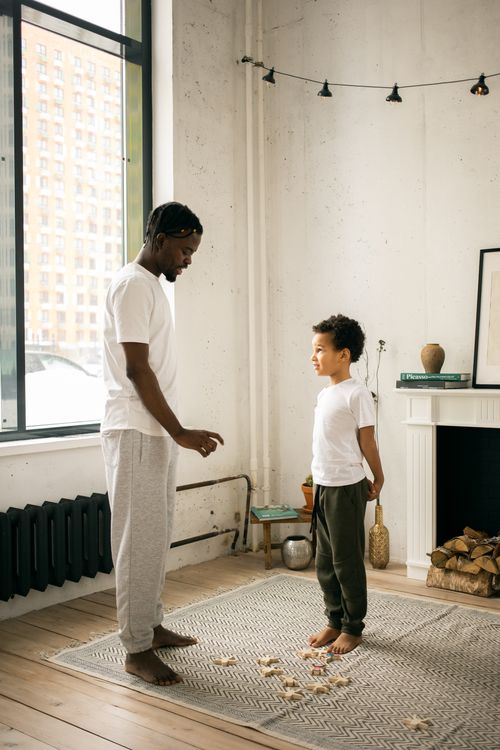 Free Side view of young barefooted African American father in casual clothes scolding little son scattered wooden toys on carpet at home Stock Photo