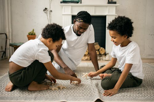 Happy young African American father in casual outfit smiling while sitting on floor and playing with wooden toys with positive twin sons during weekend at home