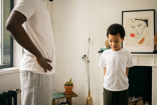 Crop unrecognizable black father standing with hands on waist and scolding confused son
