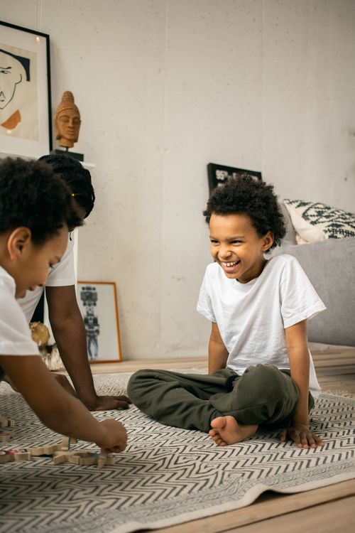 Free Cheerful young African American little brothers with curly hair in white t shirts laughing while sitting on floor and playing with wooden blocks at home Stock Photo