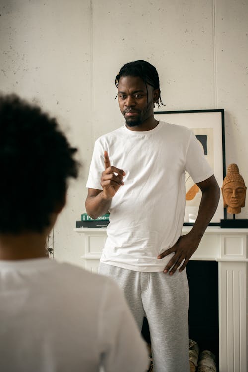 Dissatisfied young bearded African American man in white t shirt standing with hand on waist and edifying crop anonymous son at home
