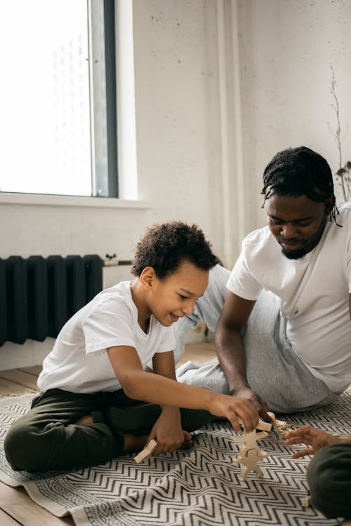 Free Pensive African American father in casual clothes sitting on floor and playing in board game with little son at home in daytime Stock Photo