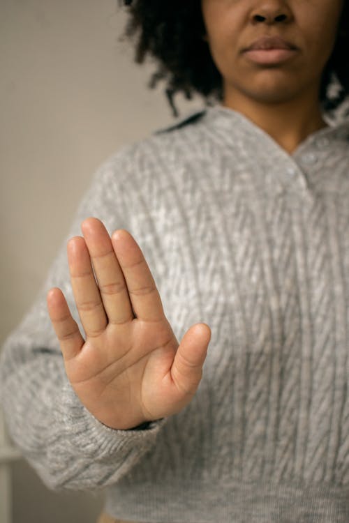 Crop anonymous African American female with curly hair in sweater showing stop gesture against light wall in room