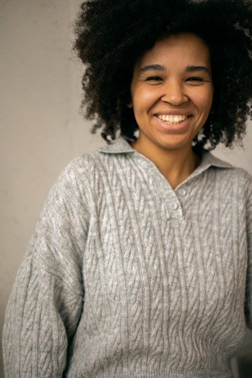 Free Crop positive African American female with short curly hair in sweater smiling and looking at camera against light wall Stock Photo