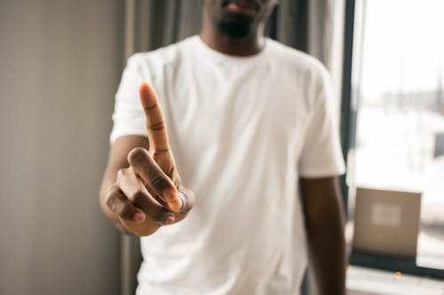 Crop anonymous African American male in white t shirt showing stop gesture with index finger pointing up in light room on blurred background