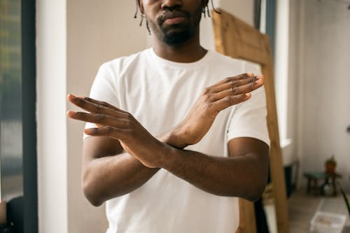 Crop unrecognizable bearded African American male in white t shirt demonstrating no gesture with hands while standing in light room