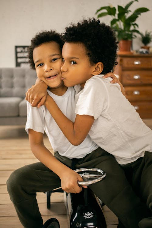 Cheerful African American brothers in casual wear embracing while sitting on toy car in light room with couch and wooden cupboard