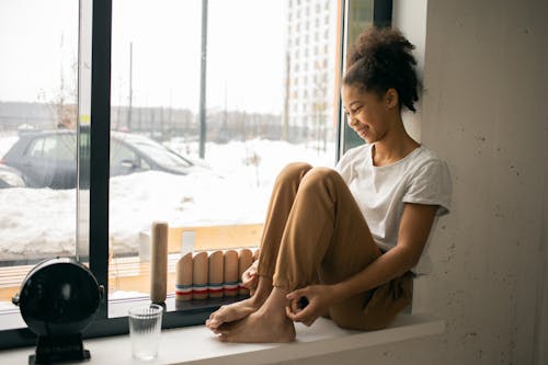 Full body side view of cheerful barefoot African American teenage girl in casual wear sitting near window in light room