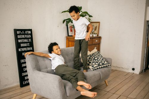 Free Full body of funny barefoot African American boys playing on comfortable couch in light room with signboard and wooden cupboard Stock Photo