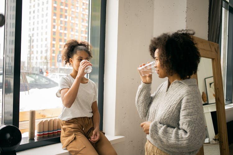 Content Black Woman With Daughter Drinking Water Near Window