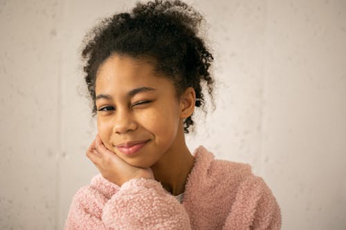 Positive African American teenage girl with Afro hairstyle wearing warm pink garment winking while looking at camera on white background