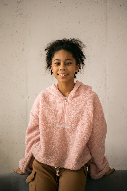 Cheerful African American teenage girl in casual clothes looking at camera while sitting on back of sofa on white background in room