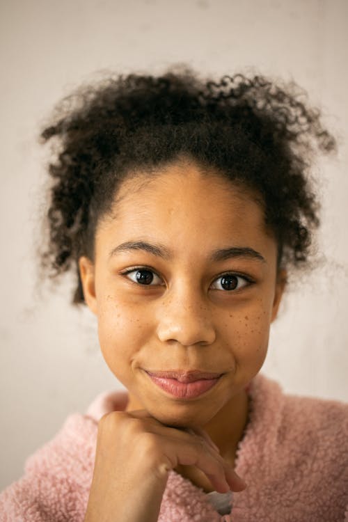 Headshot of African American teenage girl with black hair leaning on hand and looking at camera on white background in room