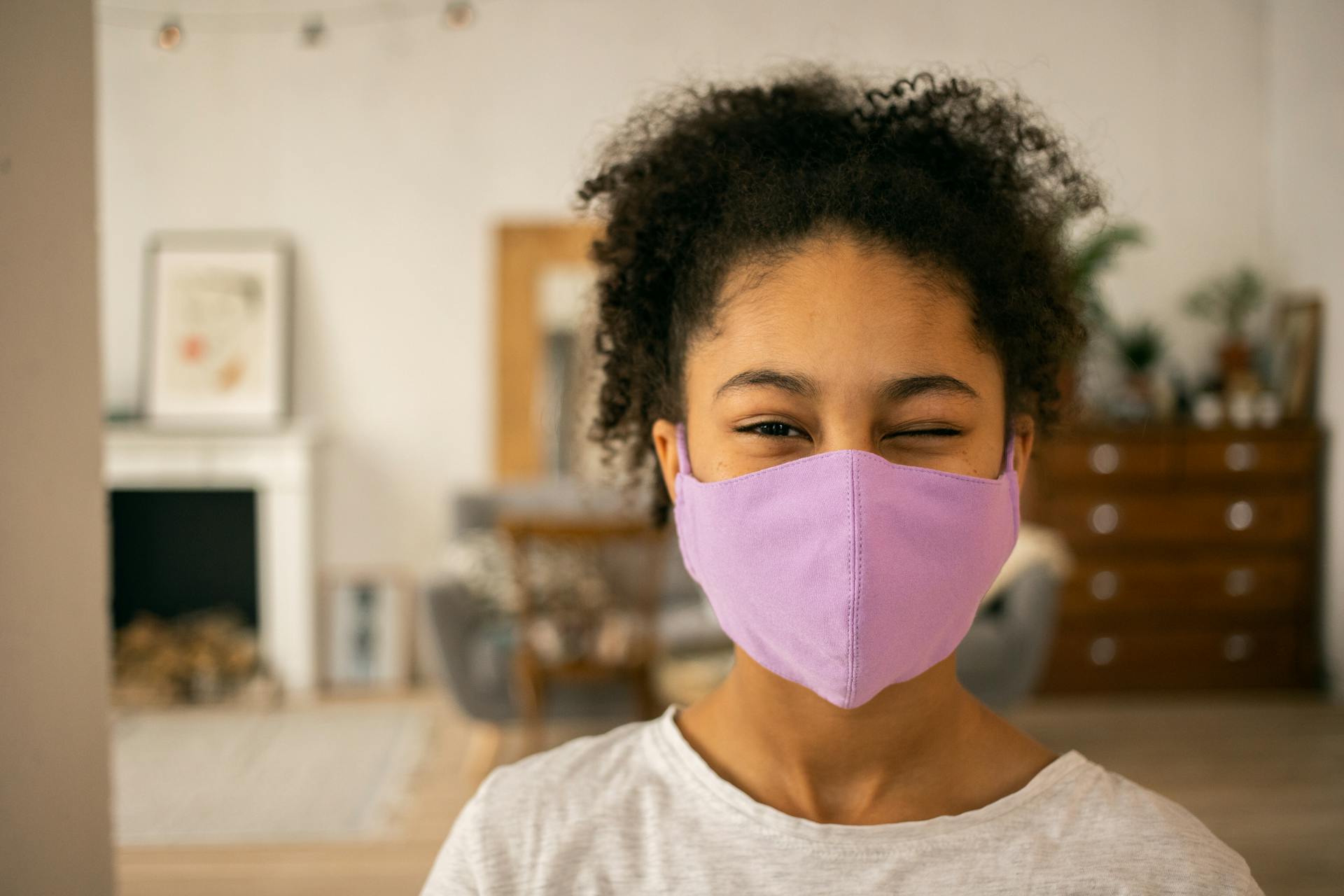 Cheerful African American teenage girl wearing protective mask winking and looking at camera on blurred background during coronavirus pandemic at home