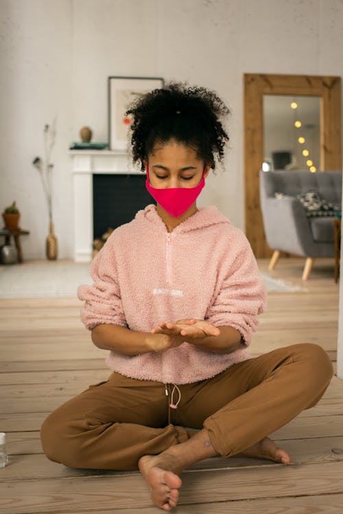 Free Full body of barefoot African American teenage girl in casual clothes wearing protective mask and sanitizer on hands sitting on wooden floor in room during pandemic Stock Photo