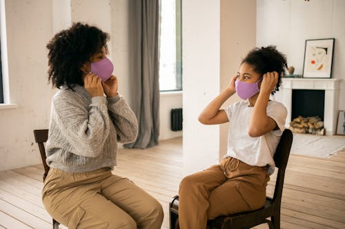 Free Side view of African American mother and daughter wearing protective masks looking at each other while sitting in room with fireplace during coronavirus pandemic Stock Photo