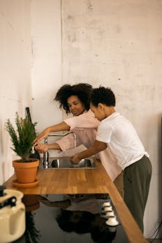 African American mother with son washing hands at sink near wooden kitchen counter with flowerpot and cooker in light apartment