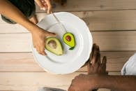 Top view of crop anonymous African American people near white plate with half of ripe avocado and sweet lollipop with similar shape