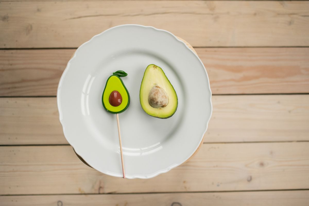 Top view half of ripe avocado near sweet lollipop with similar shape served on white plate in room with wooden floor