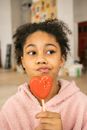 Adorable African American teenage girl with tasty sweet red gingerbread cookie on stock pouting lips and looking away in light room at home