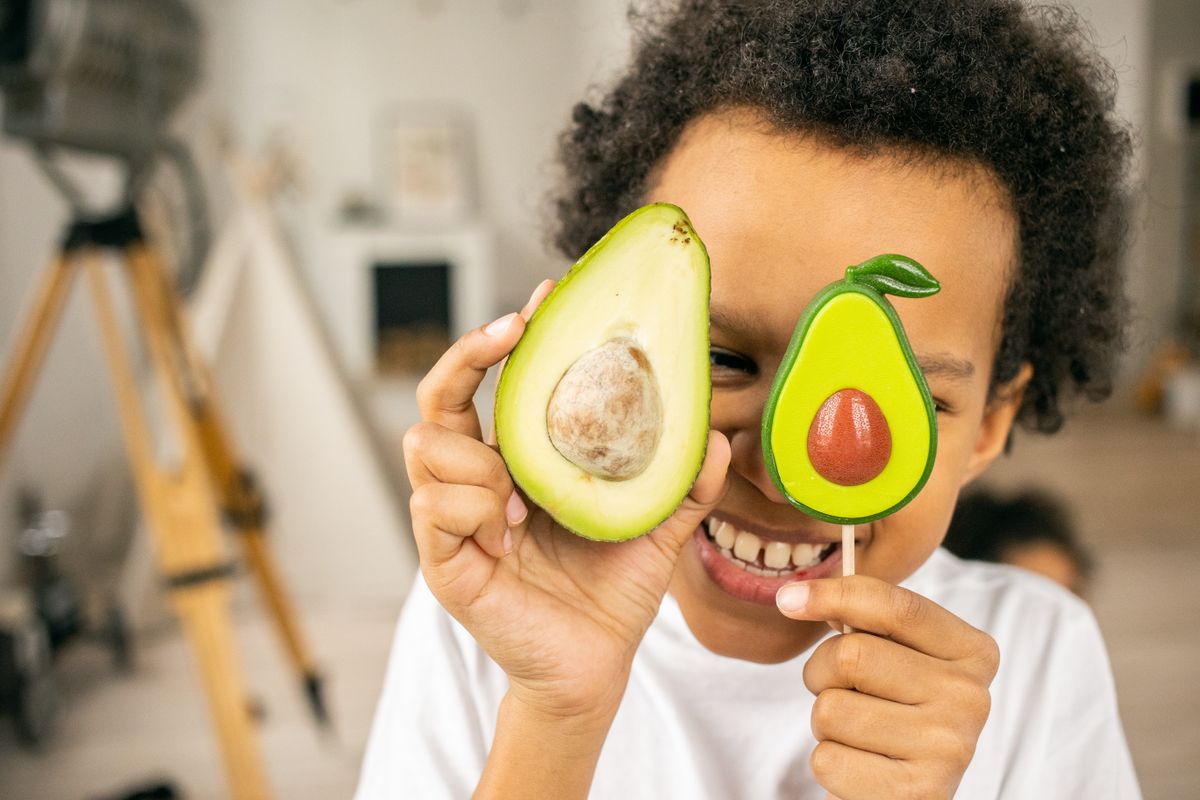 Cheerful African American boy covering face with half of ripe avocado and sweet lollipop in similar shape while standing in light room on blurred background