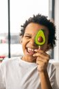 Positive African American boy in casual wear winking and covering eye with tasty lollipop in shape of avocado in light room