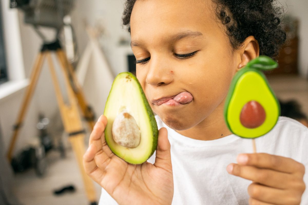 Crop African American boy with half of ripe avocado and avocado lollipop in hands while standing in room on blurred background