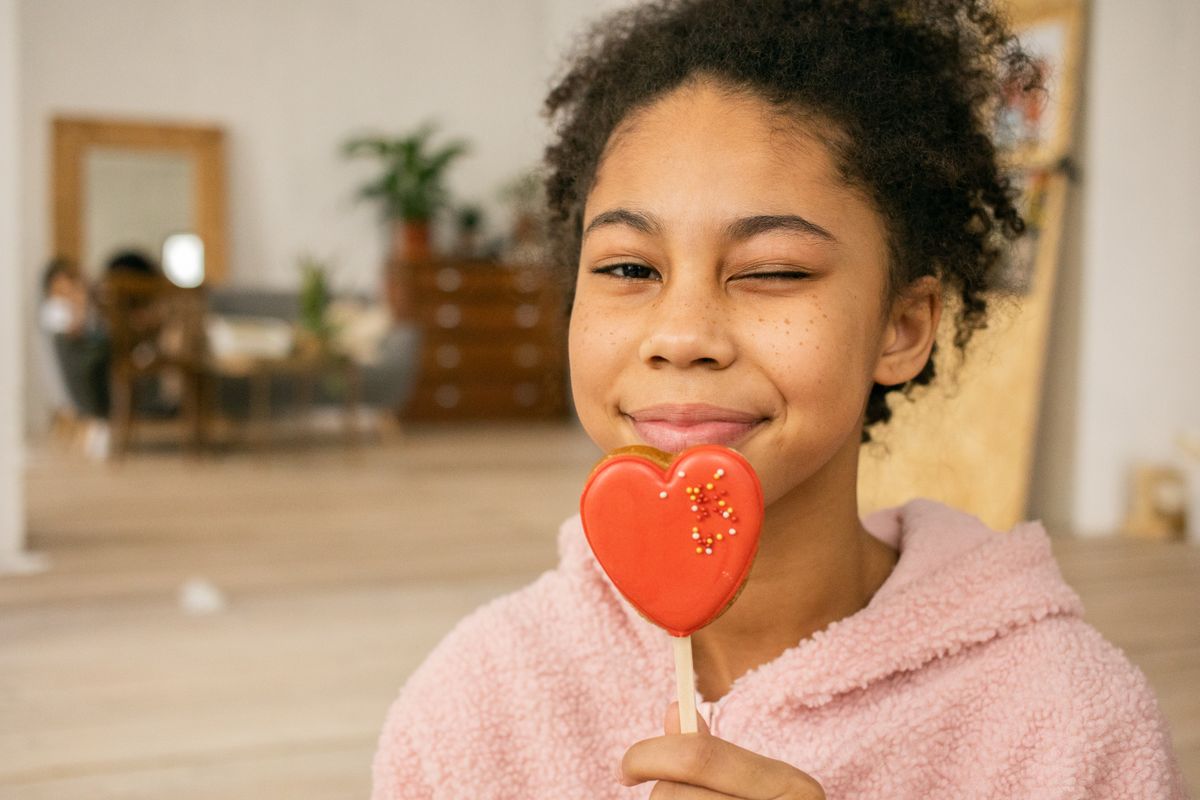 Happy African American teenage girl with sweet heart shaped gingerbread cookie on stick winking and looking at camera in light room on blurred background