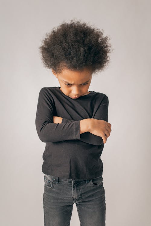 Sad African American girl with Afro hairstyle and black outfit standing with crossed arms on white background in light studio