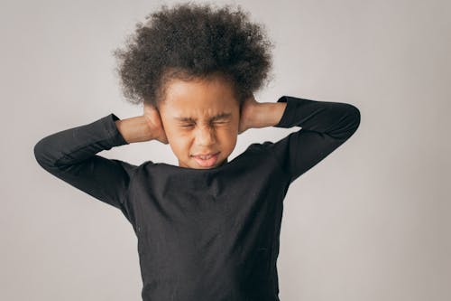 Free Unhappy African American girl with Afro hairstyle covering ears while standing on white background with closed eyes in light studio Stock Photo