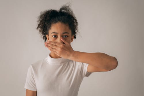 Free Concerned African American teenage girl in white outfit looking at camera while covering mouth with hand on gray background in studio Stock Photo