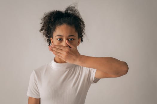 Concerned African American teenage girl in white outfit looking at camera while covering mouth with hand on gray background in studio