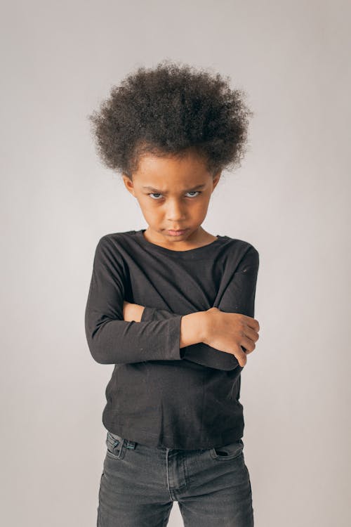 Frustrated African American girl frowning and looking at camera while standing on white background with crossed arms in light studio