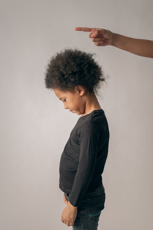 Free Side view of unhappy African American girl standing on white background near crop faceless person scolding with point up index finger Stock Photo