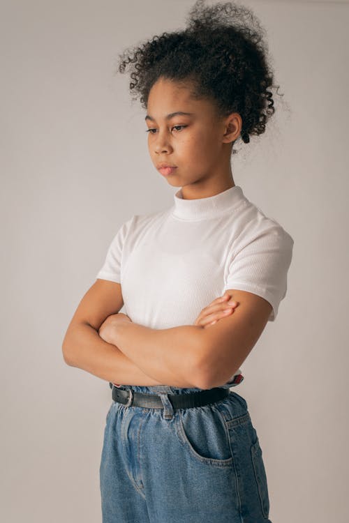 Free Sad African American teenage girl in casual clothes and black hair standing on white background with crossed arms in light room Stock Photo
