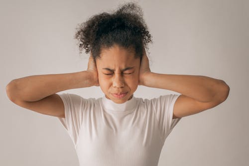 Despaired African American teenage girl with black hair and closed eyes covering ears while standing on white background in light studio