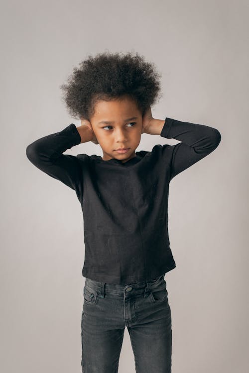 Desperate African American girl in black outfit and Afro hairstyle looking away while covering ears on white background in room