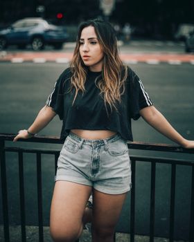 Trendy woman with dyed hair, denim shorts, and crop top leaning on a railing in urban setting.