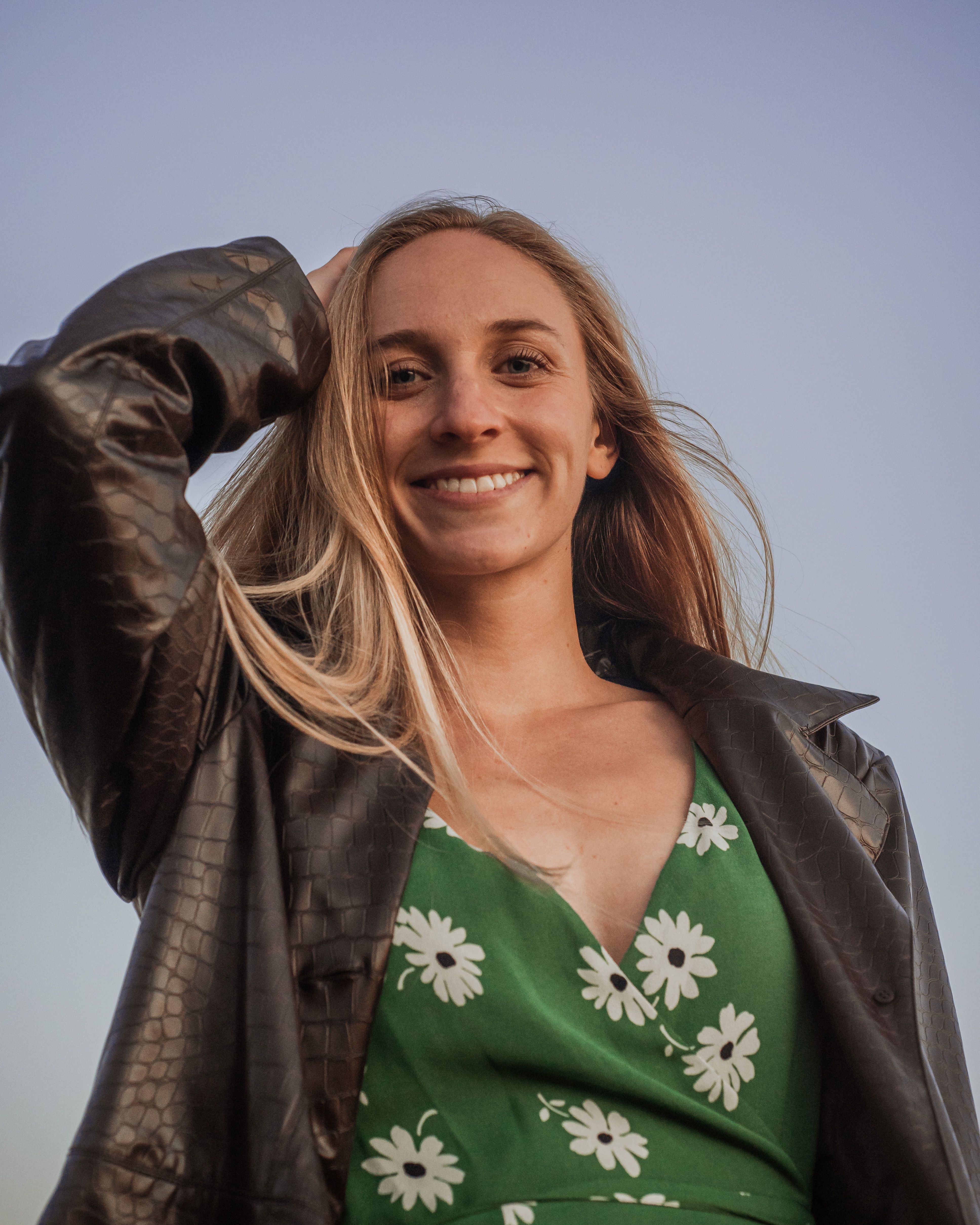 A cheerful woman in a green floral dress and leather jacket smiles warmly outdoors.