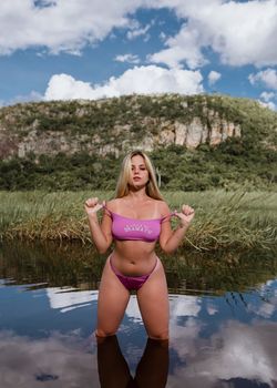 Alluring female in pink swimwear standing in water near grassy coast against cliffs and blue sky with clouds