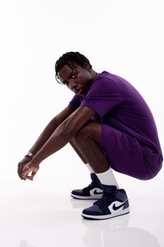 Side view of African American male in trendy sportswear squatting down and looking at camera in white studio